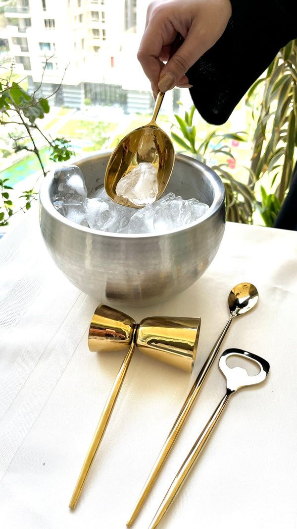 A person using a golden ladle to scoop ice from a stainless steel bowl, next to the Mádira Gilded Barware Set which includes a golden ice pick, stirrer, and bottle opener laid out on a white cloth.