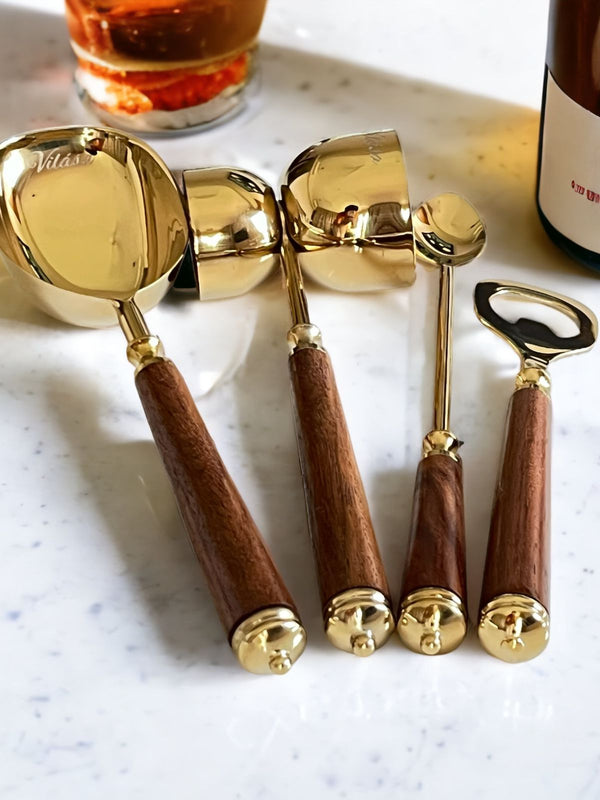 Mádira Wood Bar Tools Set including a gold drink measure, stirrer, ice pick, and bottle opener with elegant acacia wood handles on a marble countertop.