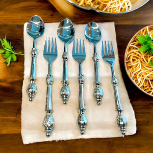 Amála Saghir 12-piece spoon and fork set displayed on a beige napkin, featuring ornate, hand-carved brass handles with a shiny finish, arranged next to a bowl of pasta on a wooden table.