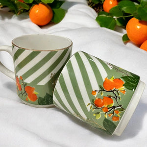 Two Gardenia Porcelain Coffee Mugs with green and white diagonal stripes, adorned with vibrant tangerine illustrations and gold detailing, displayed on a white fabric with fresh tangerines in the background.