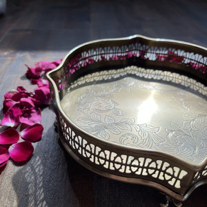 A close-up of the Názm Petal solid brass tray with delicate floral engravings and ornate cut-out details along the edge, creating a vintage and elegant aesthetic. The tray is surrounded by rose petals, adding a touch of luxury to the scene.
