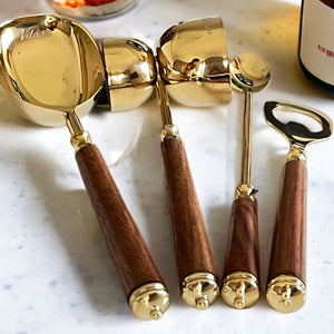 Mádira Wood Bar Tools Set including a gold drink measure, stirrer, ice pick, and bottle opener with elegant acacia wood handles on a marble countertop.