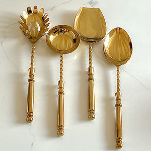 An elegant set of four Amála serving spoons with a shiny gold finish, featuring distinct designs: a deep ladle, a flat spoon, a solid spoon, and a unique spaghetti/noodle spoon, all with ornate twisted handles displayed on a marble surface.