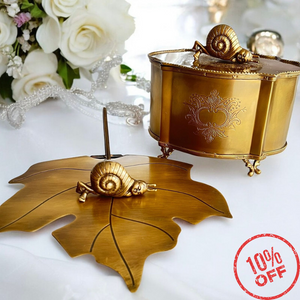 Golden brass Zen storage box and Grace napkin holder, both adorned with snail designs, set against a backdrop of white flowers and beads. The storage box features ornate legs and a lid handle, with a '10% OFF' label, emphasizing elegance and a slow-paced lifestyle theme.